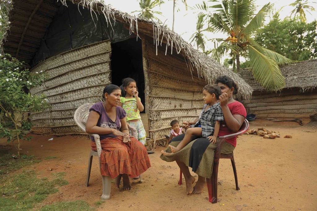 There are Sri Lankan women and children sitting in chairs outside of a hut/home talking.