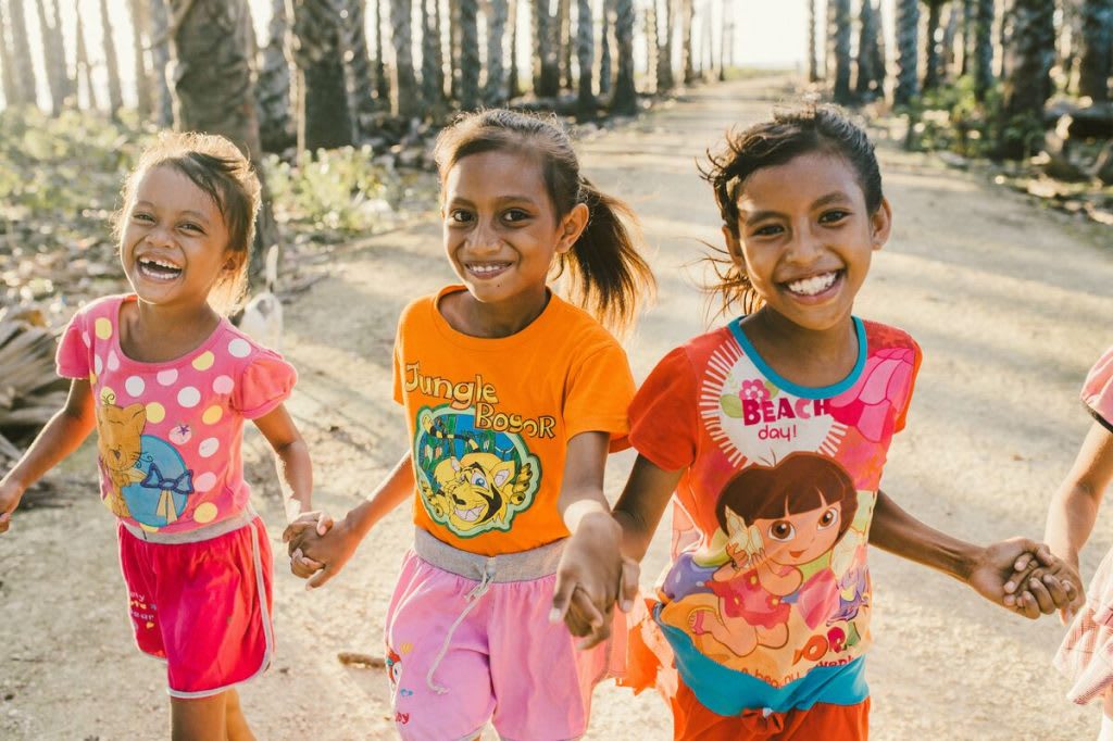 Three little girls are running on a trail together laughing. They are wearing bright pink and orange clothing.