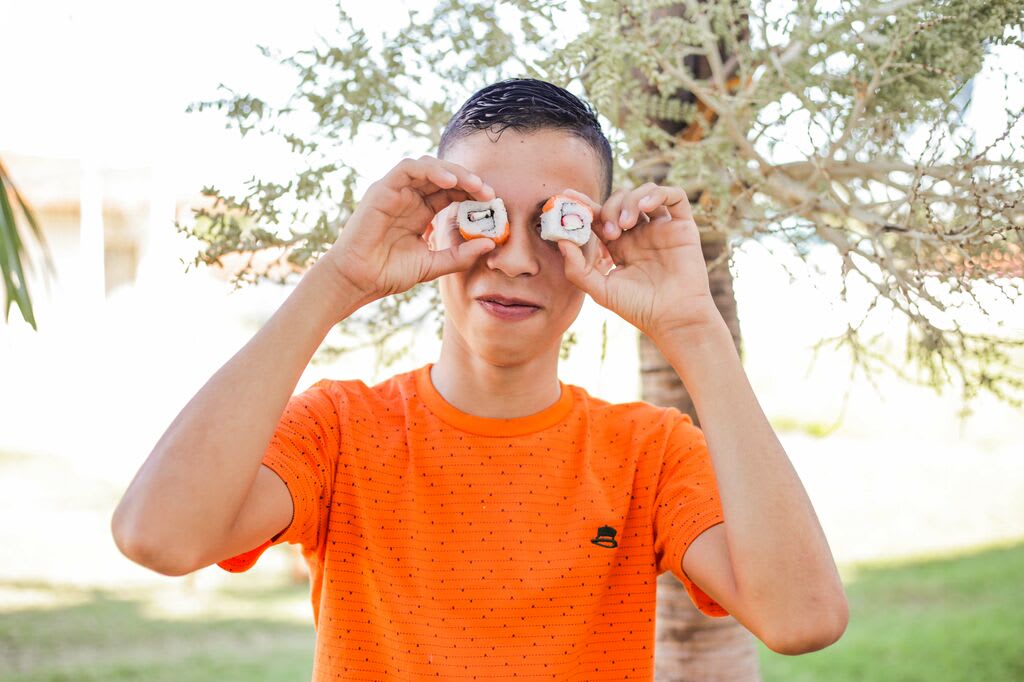 A teen boy wearing an orange t-shirt hold sushi rolls to his eyes like glasses.