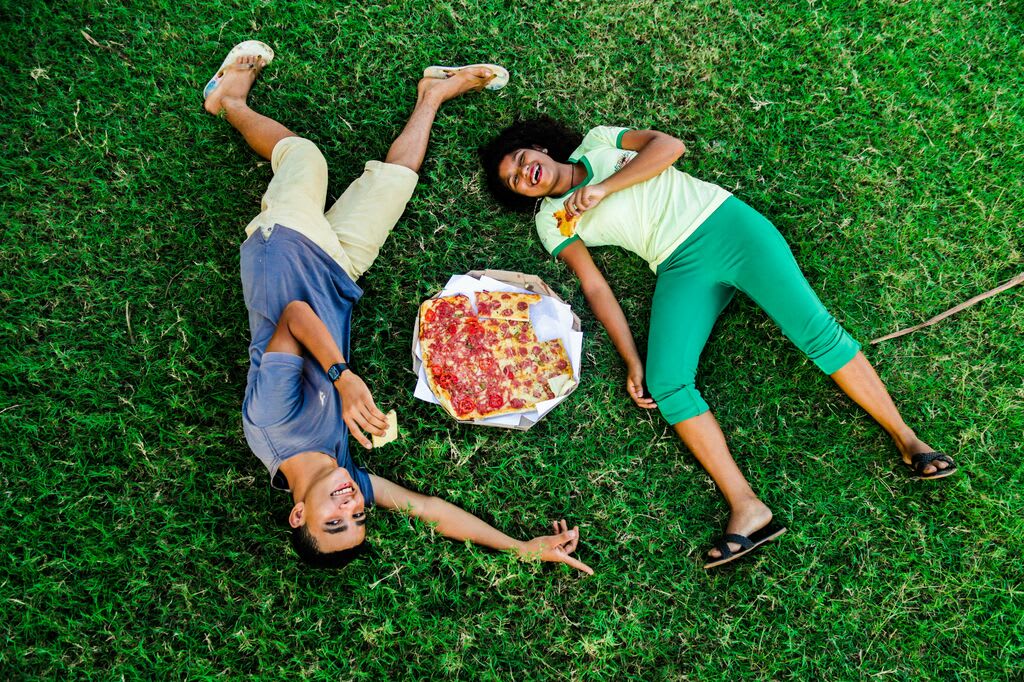 A boy and a girl lie in the grass around a pepperoni pizza.