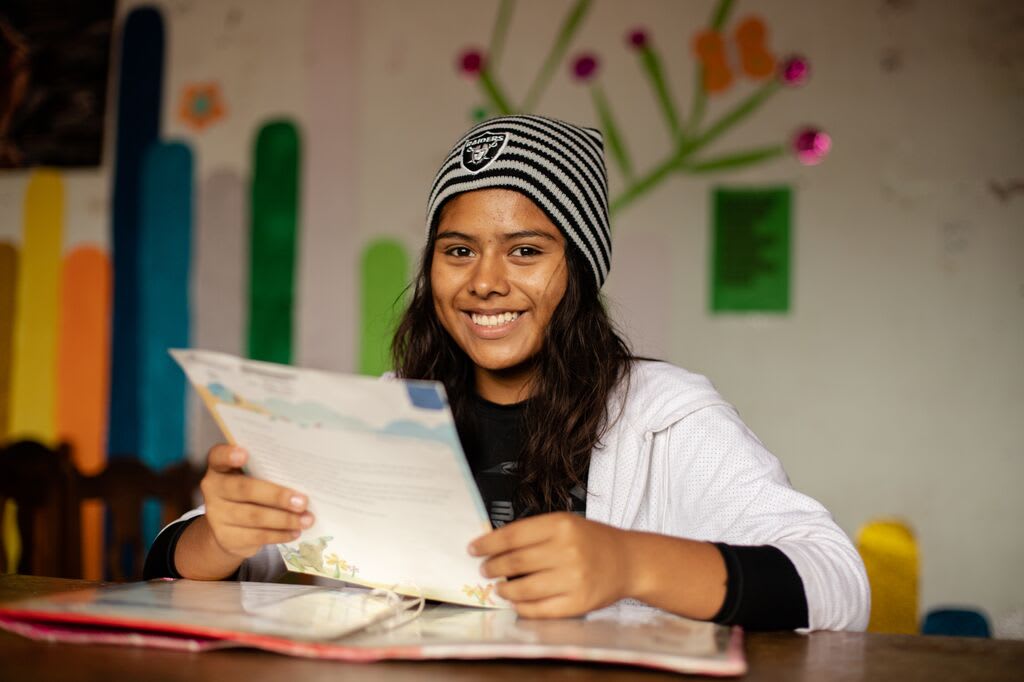 Girl smiles and looks at the camera as she reads a letter from her sponsor. She's wearing a toque.