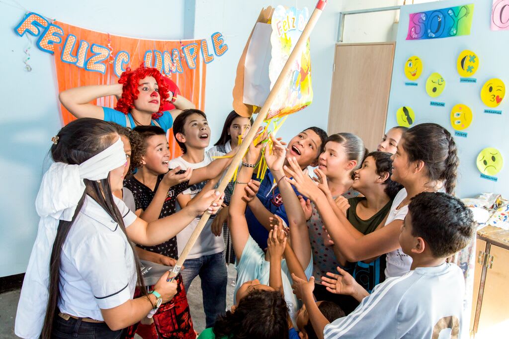Girl is blindfolded hitting the pinata that says "Feliz Cumple" . There is a teacher dressed at a clown in the back looking excited.