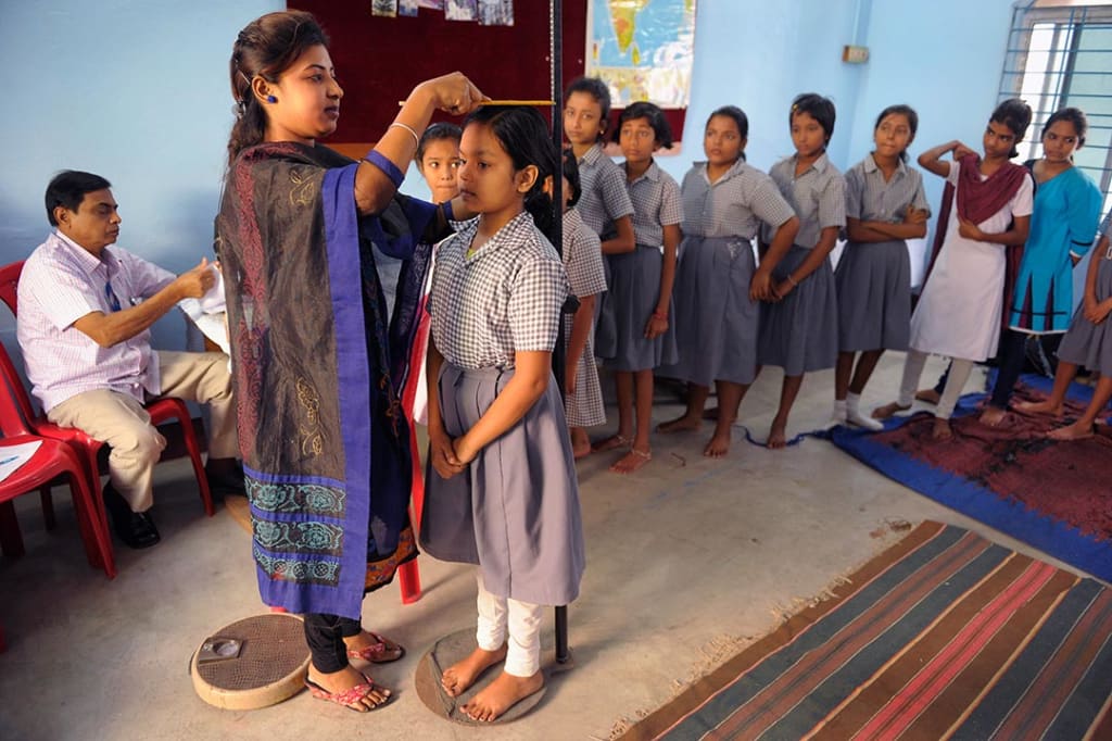 An adult women measures the height of a younger girl. They both wear traditional, Indian clothing