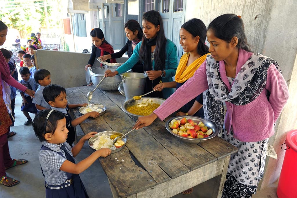 children line up by a table and hold out plates to recieve food for lunch.