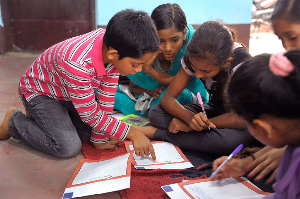 Indian children kneel around their work books and talk about their assignment.