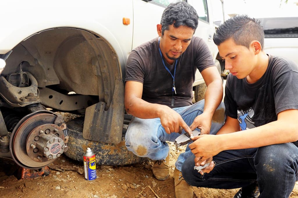 A male mentor shows a teenage boy how to work brakes on a truck.
