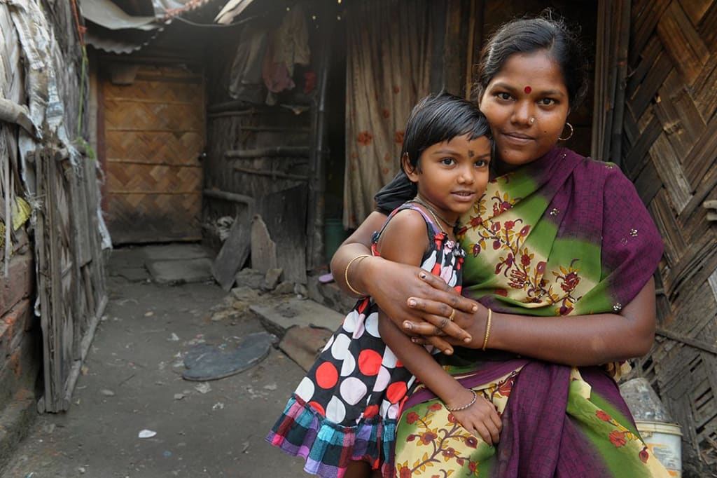 A young Indian girl is embraced by her mother. They sit in front there home which appears to be in a slum.