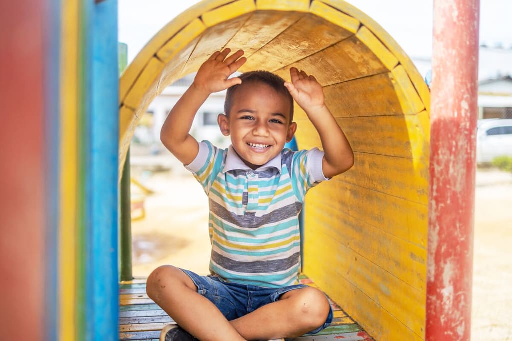 A boy sits in a yellow playground tunnel and smiles. He is learning at his centre how much Jesus loves him and the other children!
