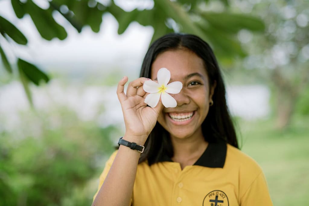 A girl in a yellow shirt holds up a white flower to her face and smiles.