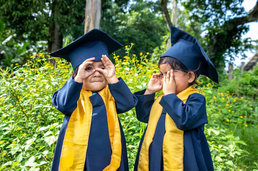 Two young children wear graduation gowns and caps and hold their hands up to their eyes.