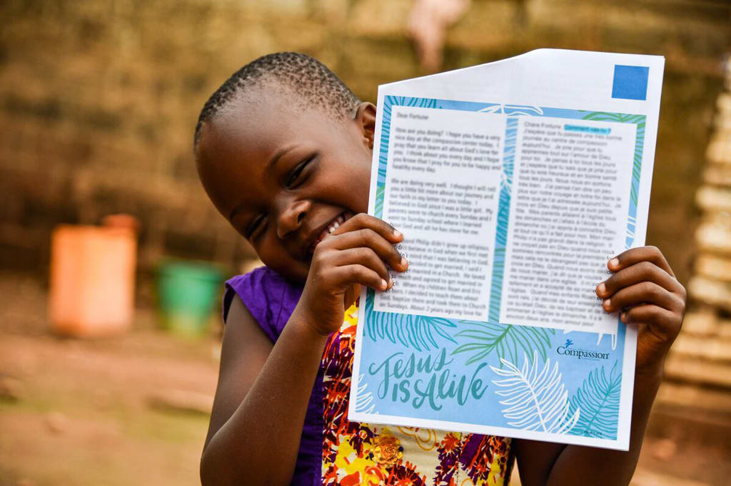 A young girl in a colourful dress holds up a letter and smiles.