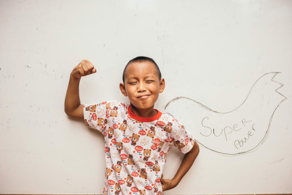 A little boy in a patterned shirt flexes his bicep and stands in front of a drawing of a cape that says "Super Power"