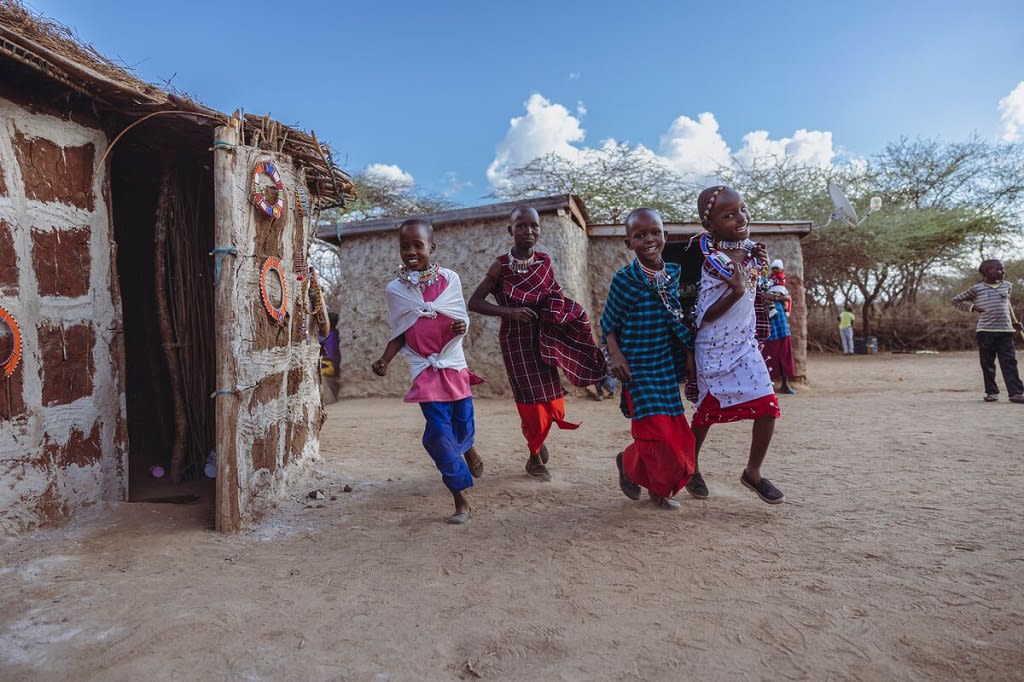 Four children dressed in traditional clothing run and laugh together besides their homes.