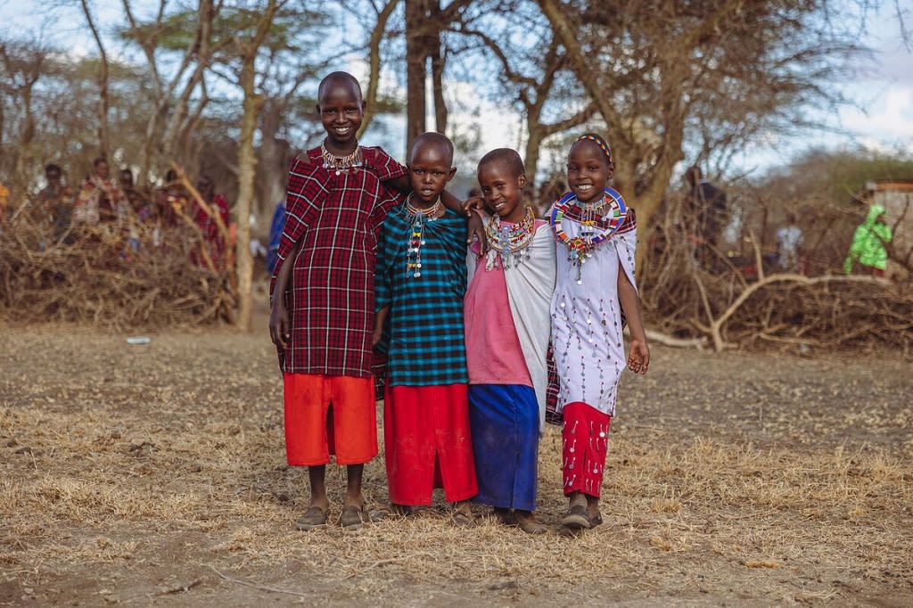 Four children stand in their traditional clothing while smiling in front of some trees.
