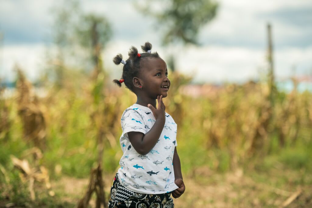 Little girl smiles off into the distance in a field. At her Compassion centre, she learns Jesus loves and protects her.
