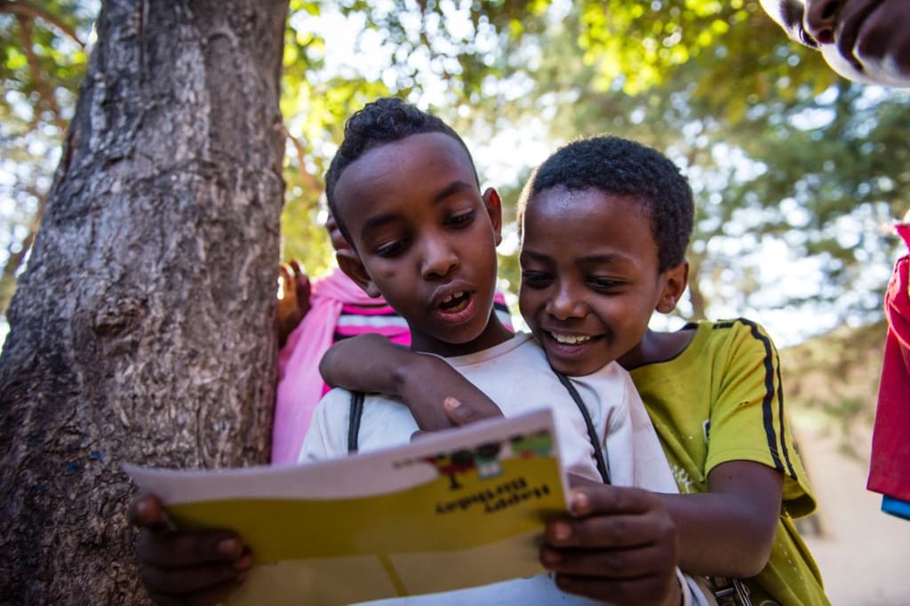 Two boys smile while reading a letter. They are standing by a big tree.