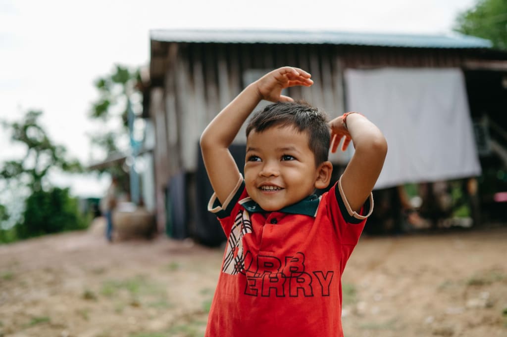 A child throws his hands up in the air in joy. He is wearing a red shirt and stands in front of a farm building.