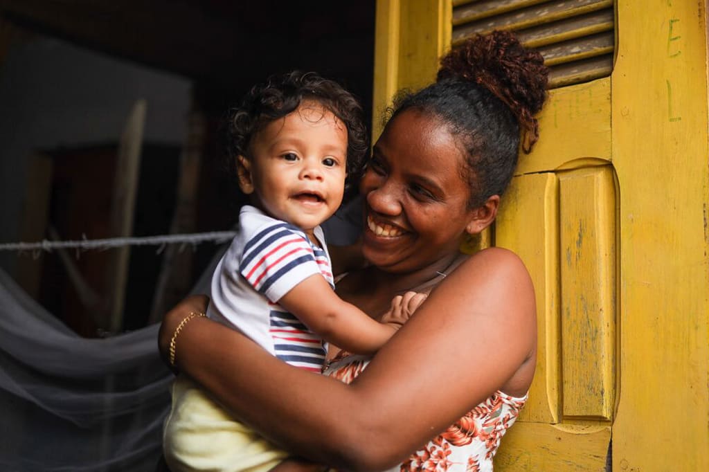 A woman smiles at a baby in her arms by a yellow door.