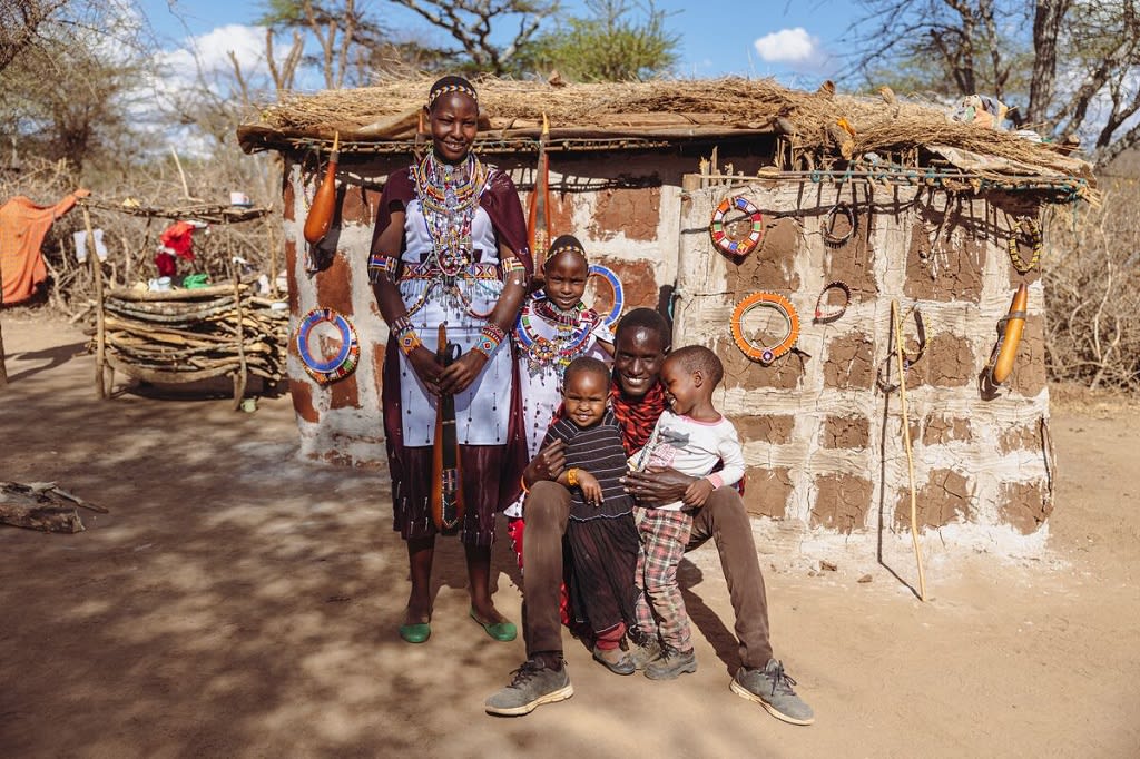 Pesi, her mother, father, and her siblings Enock and Tasha are sitting outside their home. Behind them a wall decorated with beaded jewelry. Pesi and her mother are wearing traditional clothing.