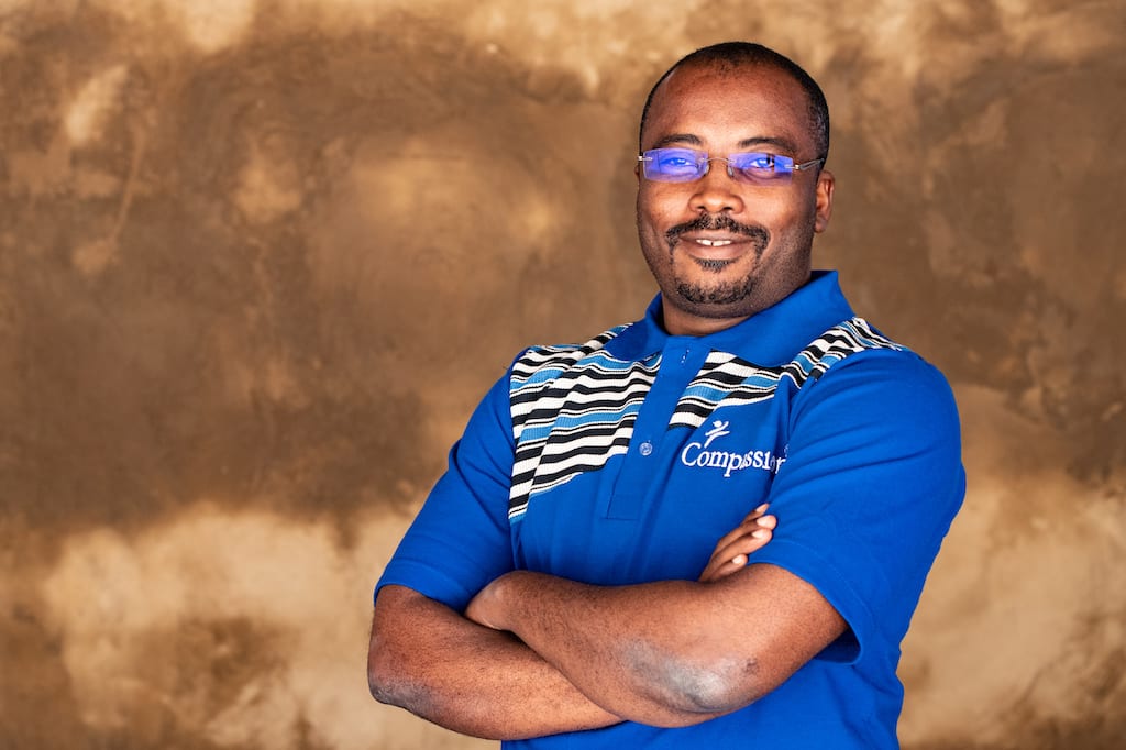Dr. Issaka Kiemtore is wearing a blue shirt with gray and black stripes and the Compassion logo on the front. He is standing in front of a brown wall with his arms crossed.