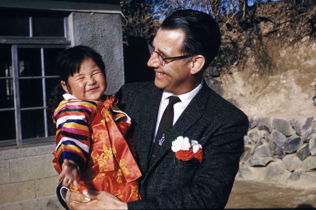 Rev. Swanson is wearing a suit and tie and carrying a Korean toddler in his arms. They are both smiling.