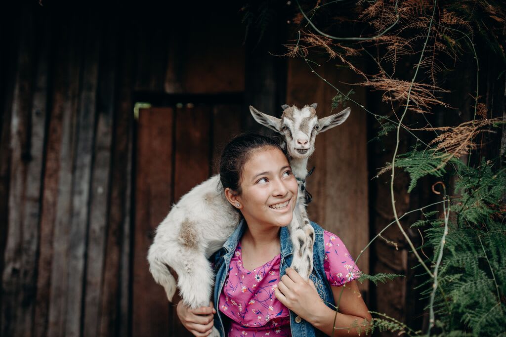 Girl wearing pink shirt carries a goat around her shoulders.