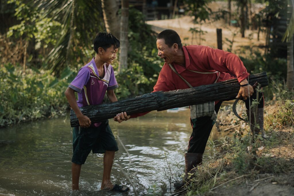 Oosamai and his son, Gungamae-ou lift a log out of the river together while laughing.