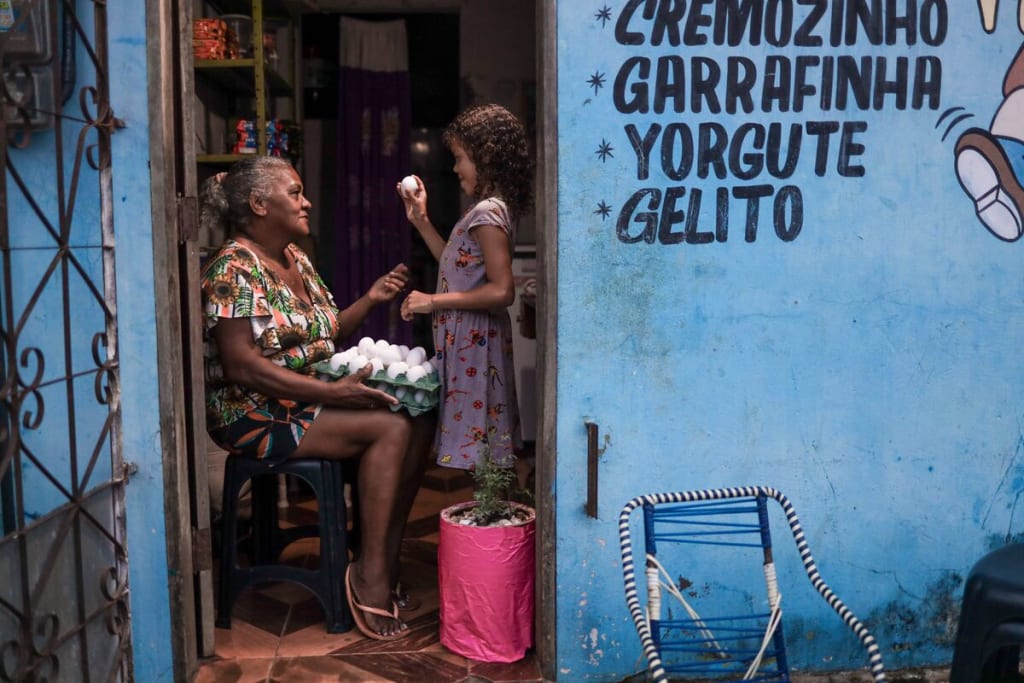 A woman sits with a carton of eggs on her lap. A young girl holds one up in front of her.