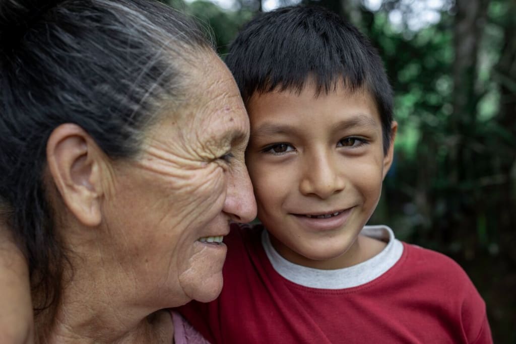 A woman smiles with her face near to a young boy in a red shirt.
