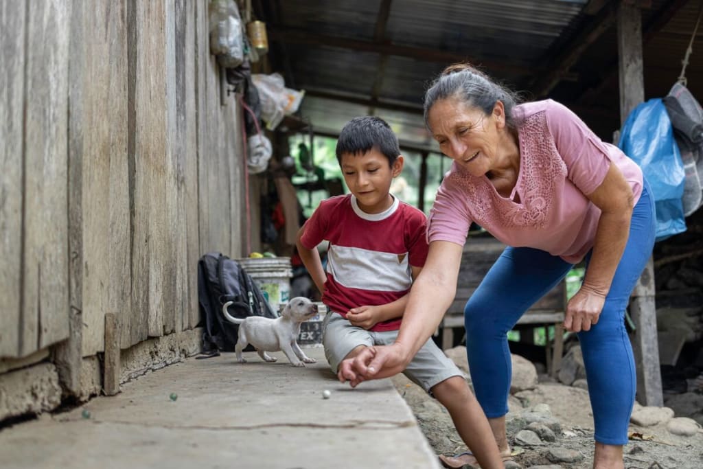 A woman in a pink shirt and jeans plays marbles with a young boy in a red shirt and shorts.
