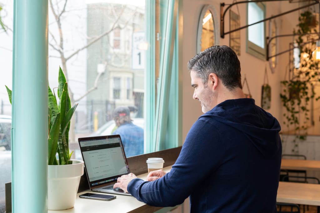 A man in a navy sweater types on his laptop in front of a window