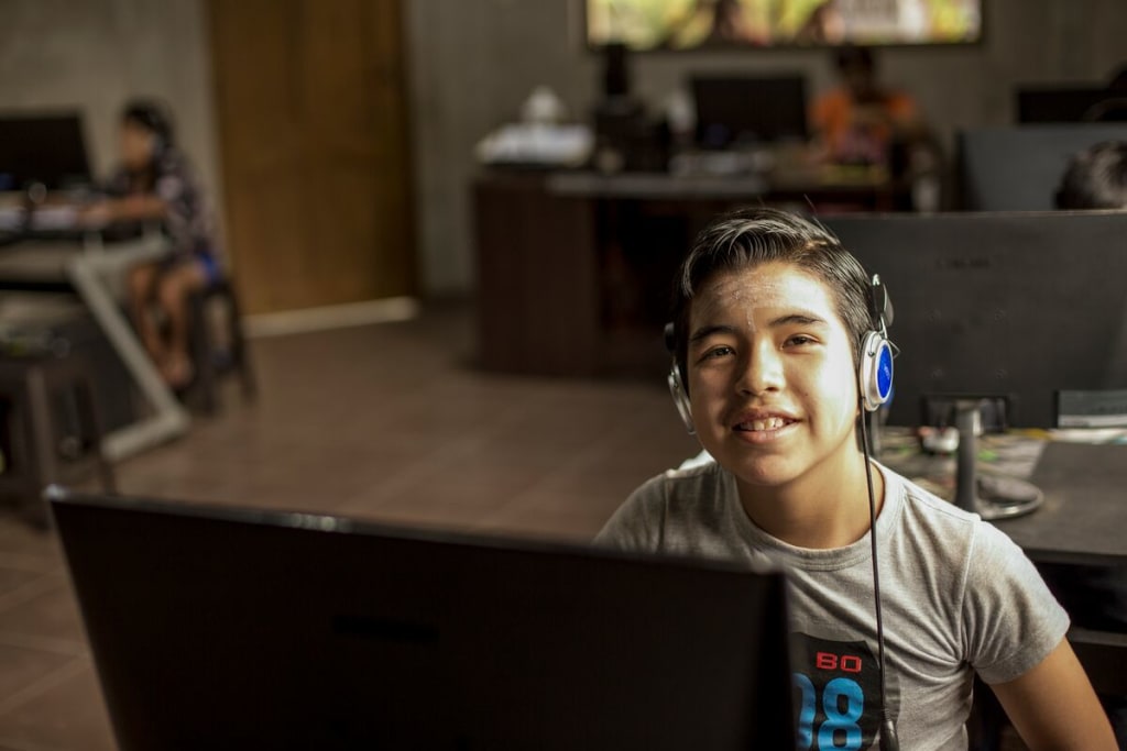 A boy sits with blue earphones on, smiling over a computer monitor