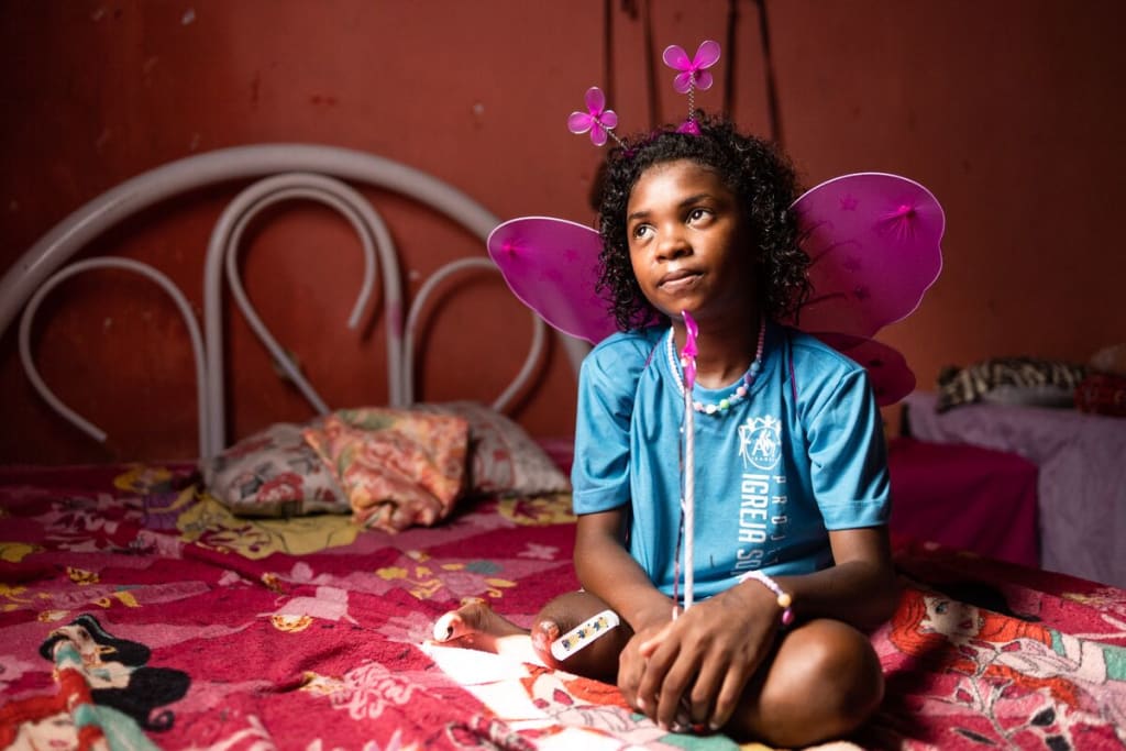A girl in a blue shirt sits on a bed in a pink room