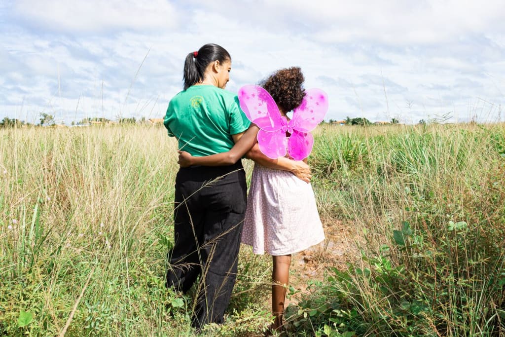 A girl in butterfly wings and a woman in a green shirt and black pants stand in a field with their backs to the camera
