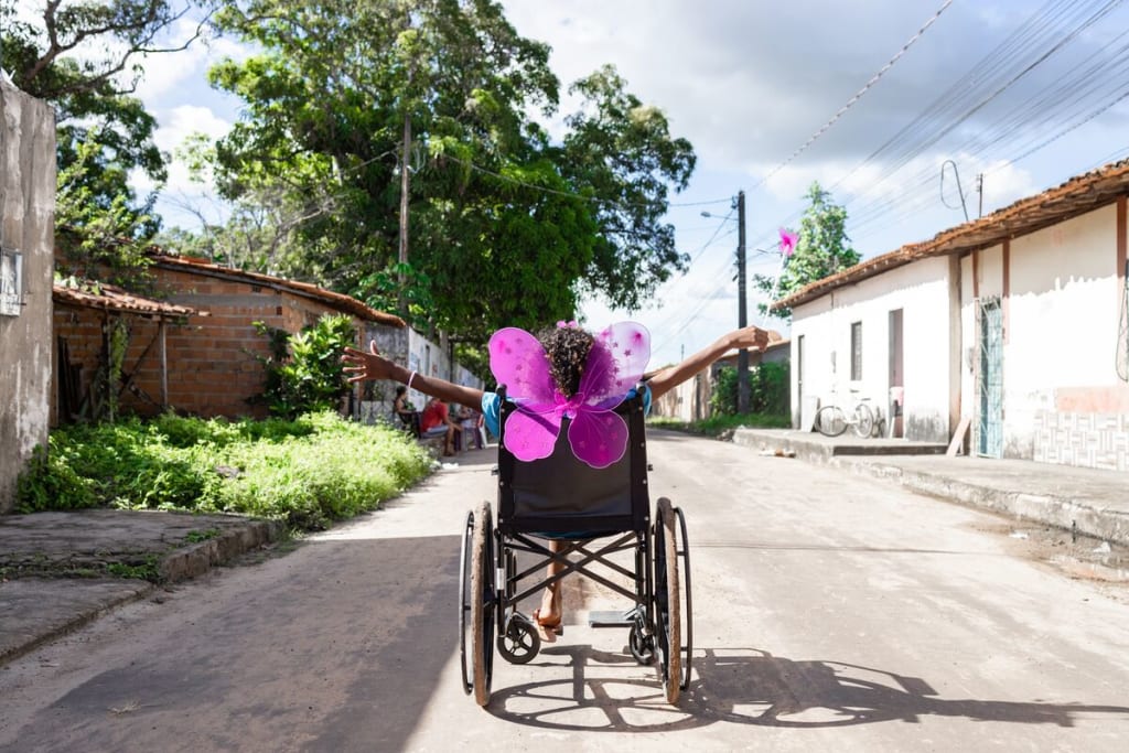 A girl in butterfly wings sits in a wheelchair with her hands outstretched
