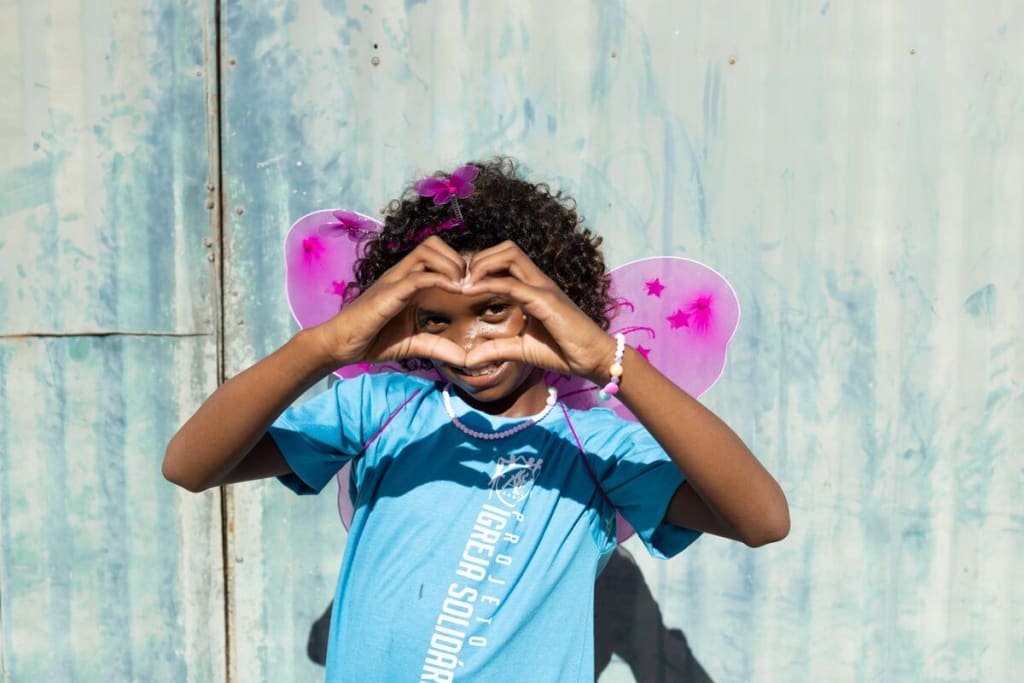 A girl in a blue shirt holds her hands in a heart shape and looks through them at the camera