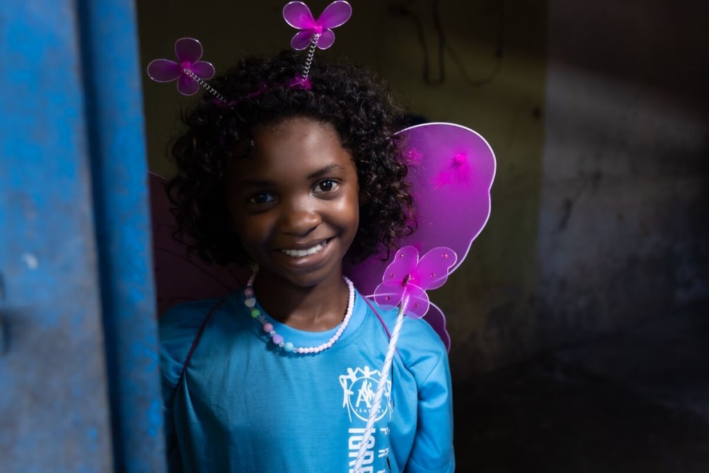 A girl smiles at the camera. She wears a blue shirt and pink wings
