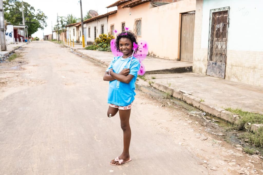 A girl in a blue shirt and pink wings stands in the street on one leg.