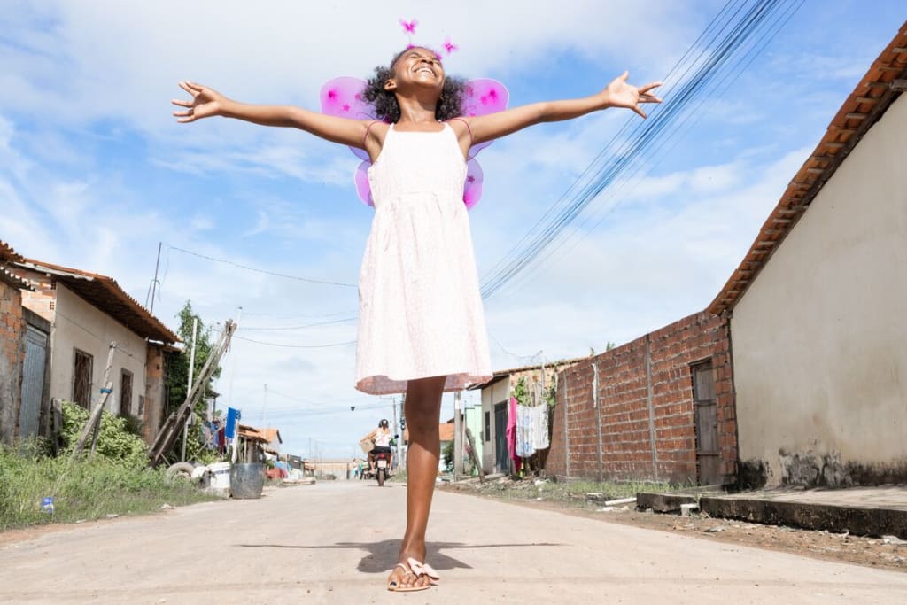 A girl in a pink dress and pink wings stands on one leg with her arms stretched wide to the sky