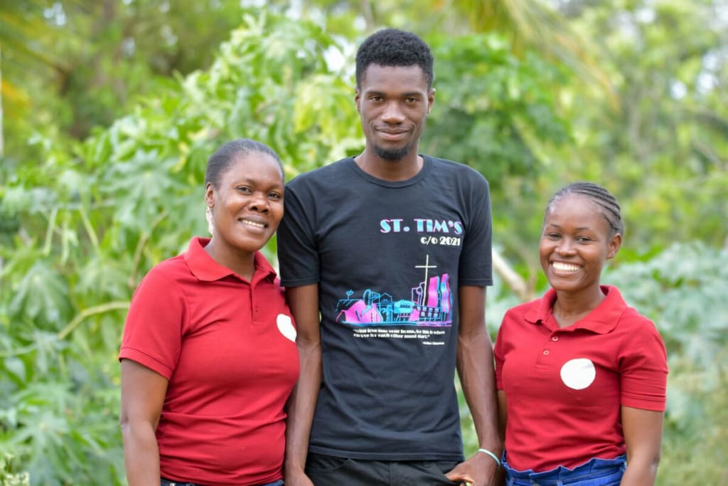 A young man in black stands with two women in red.