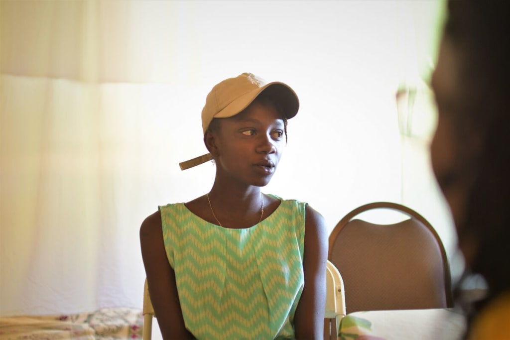 A girl in a green and yellow top and a beige baseball cap sits talking