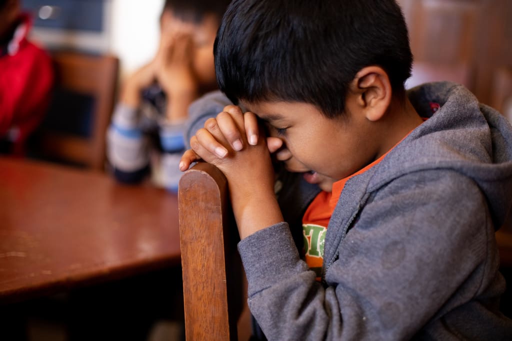 A little boy prays while sitting in a chair