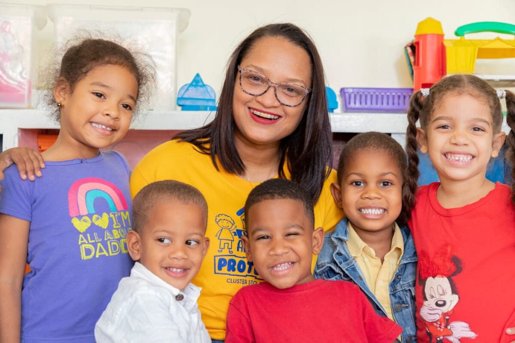 A woman in a yellow shirt sits with a group of children and smiles