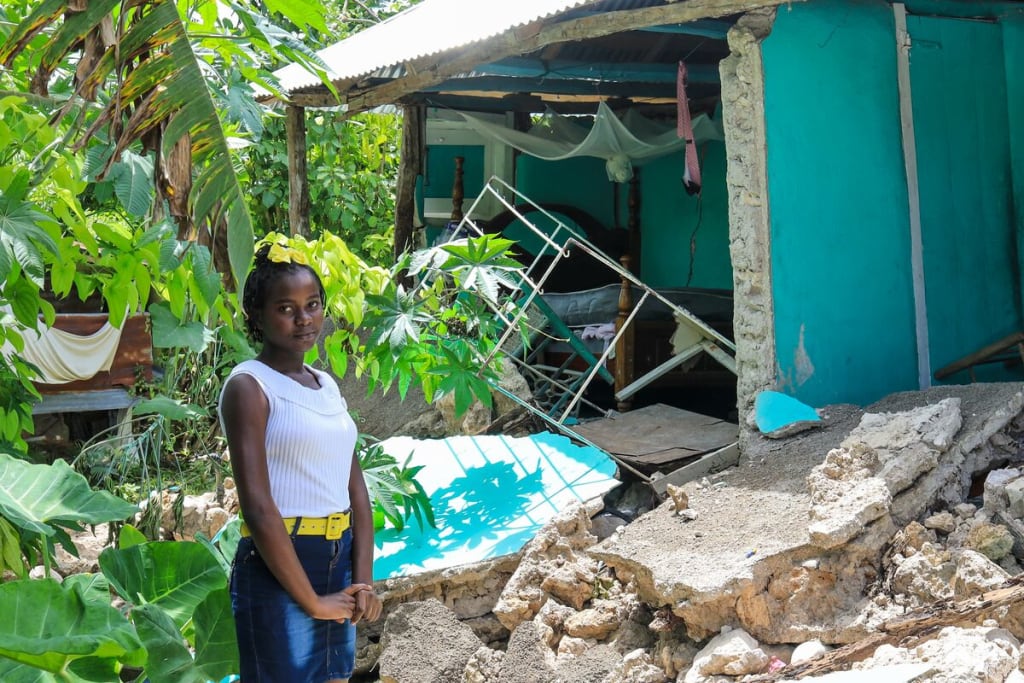 A girl in a white top and jean skirt stands in front of a home that has been damaged