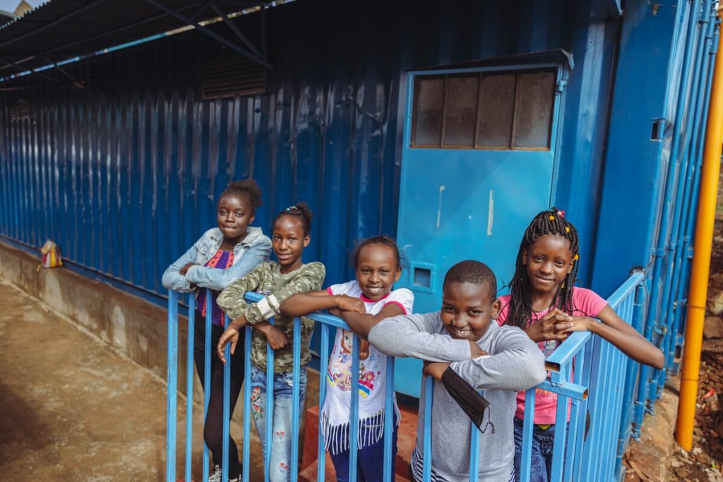 A group of children stand in front of a blue building and smile