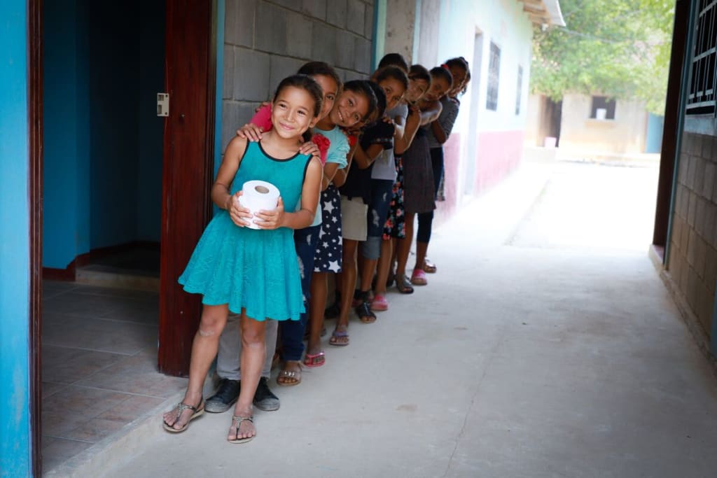 A row of children stand outside washrooms. They are smiling and holding rolls of toilet paper.