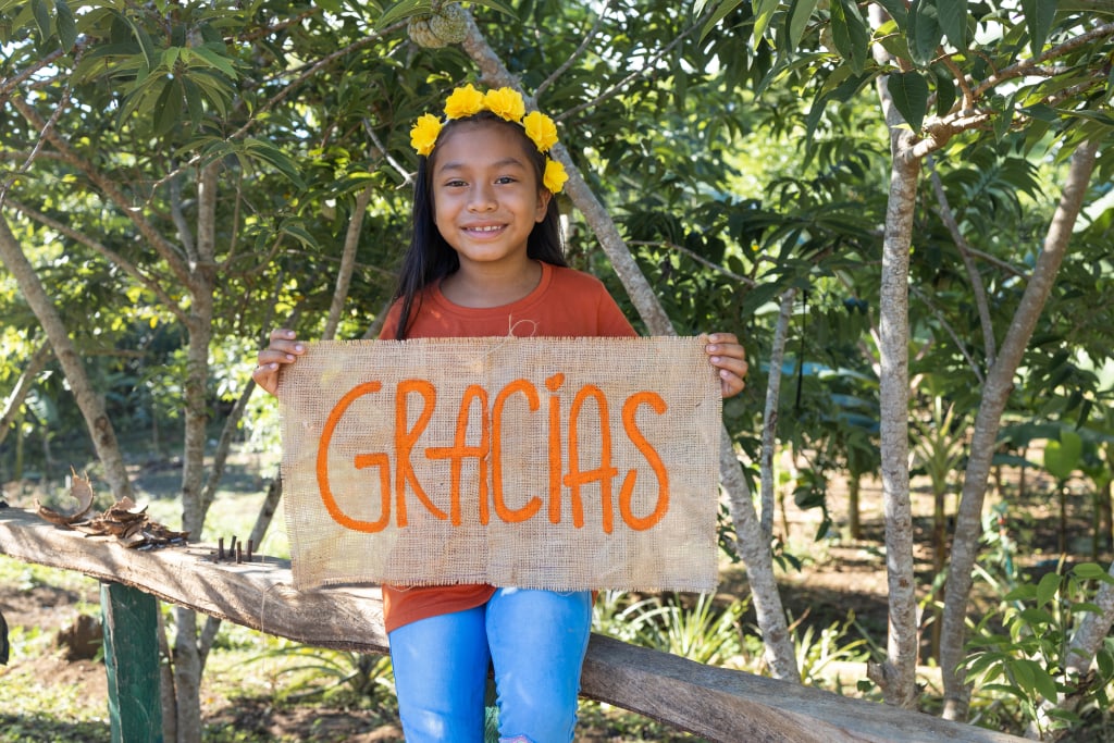 Maira is wearing an orange shirt with jeans. She is sitting on a fence around her family's garden and is holding a sign that says “thank you”.