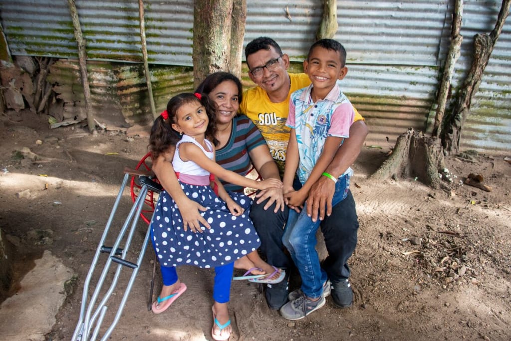 Luis is wearing a blue and white patterned shirt with pink sleeves and jeans. He is sit ting outside his home on his father's lap. Next to them are Luis' mother and sister. There are crutches to the side.