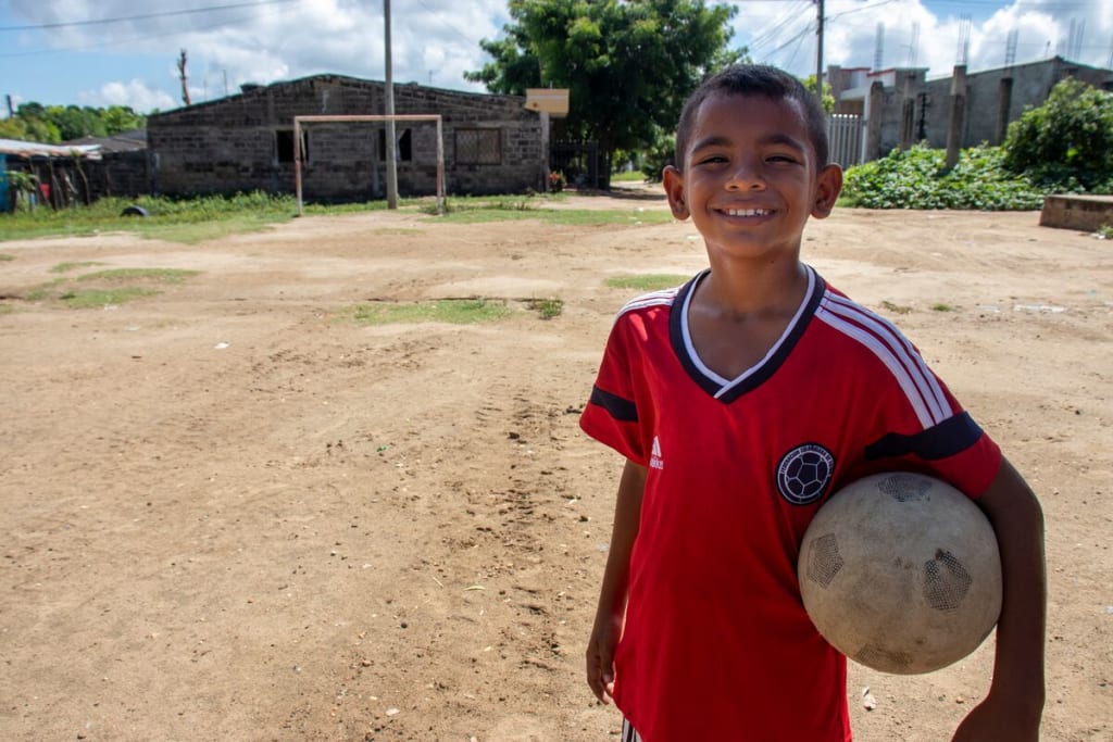 Luis is wearing a red shirt. He is outside on a soccer field and is holding a soccer ball under his arm.