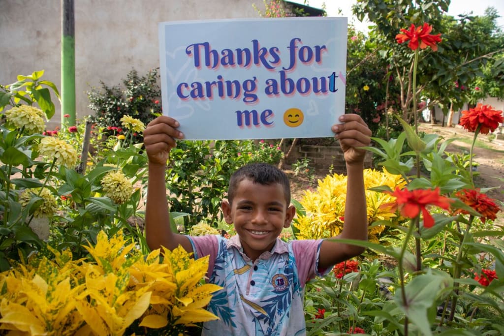Luis is wearing a blue and white patterned shirt with pink sleeves and jeans. He is standing outside surrounded by red and yellow flowers. He is holding up a sign that says, "Thanks for caring about me."